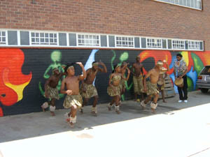 A group of black dancers are performing energetically in front of brightly coloured painted murals. On the right, a black man, wearing an elaborate beaded collar, is accompanying them on a drum.